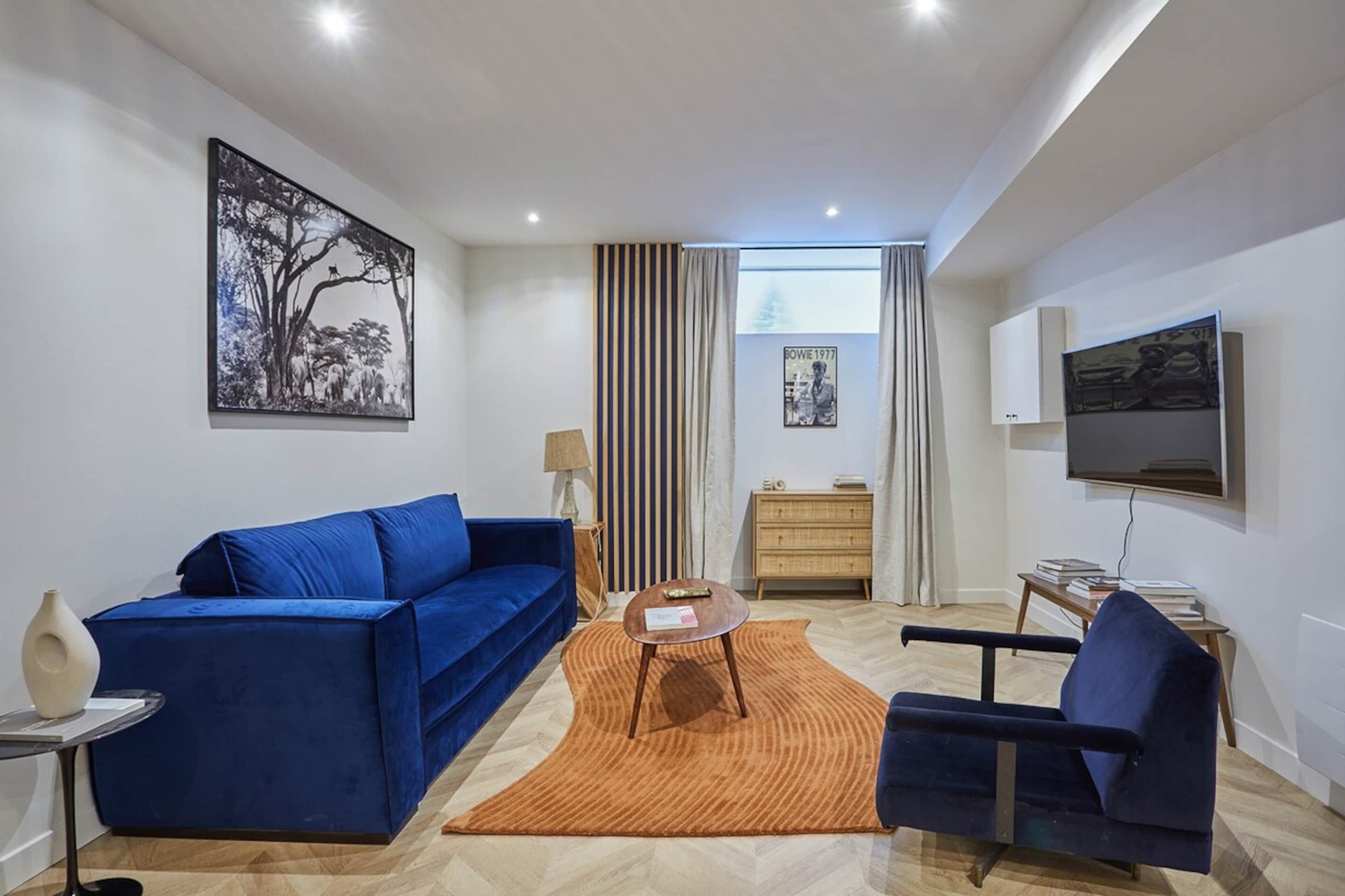 A modern living room with a blue sofa, a small round table, a blue armchair, a wooden cabinet, and a mounted TV.