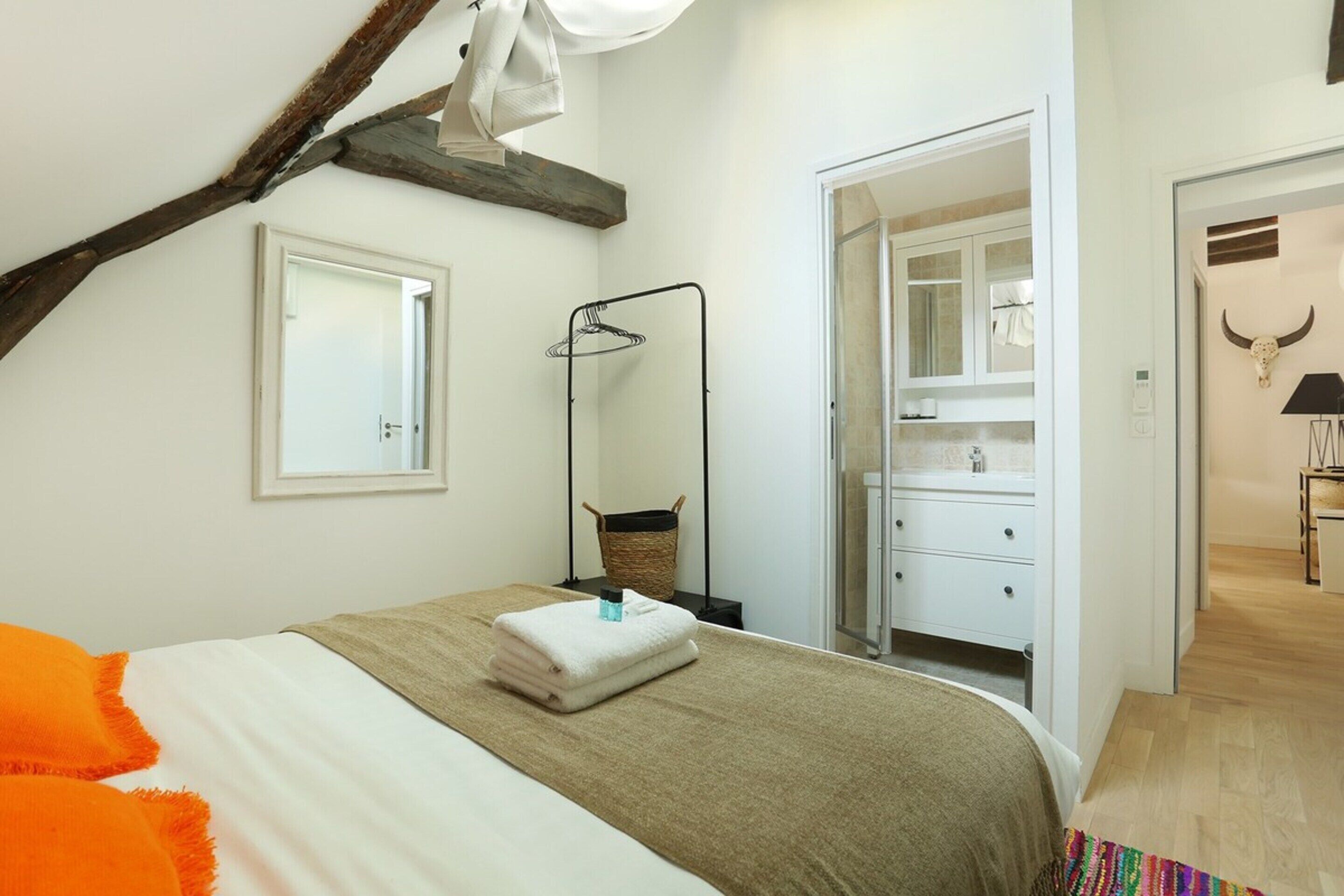 A bedroom with a bed, a wooden beam, a mirror, a hanging rack, and a bathroom visible through an open door.