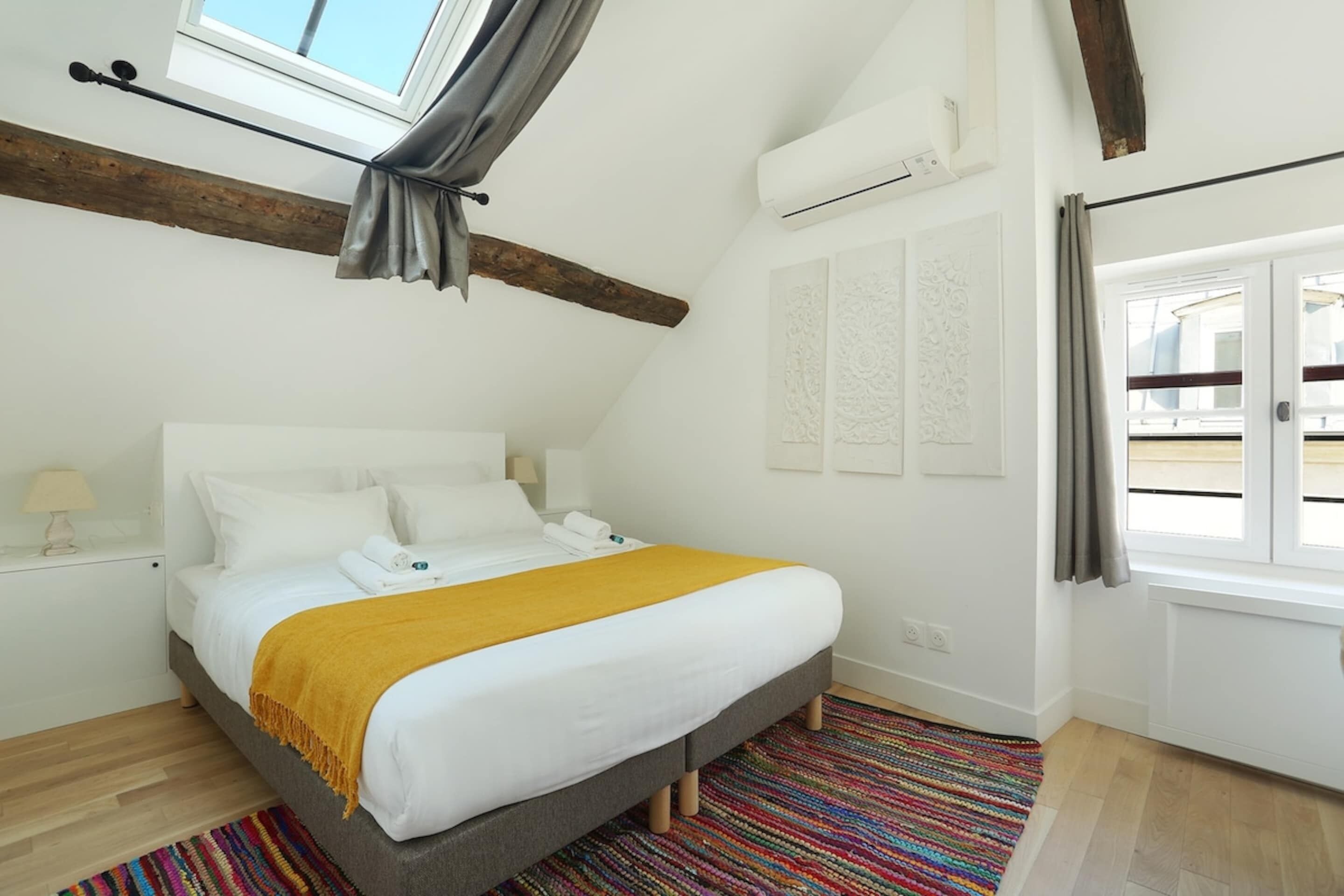 A bedroom with a bed, a window with curtains, a wooden beam ceiling, and a colorful rug.