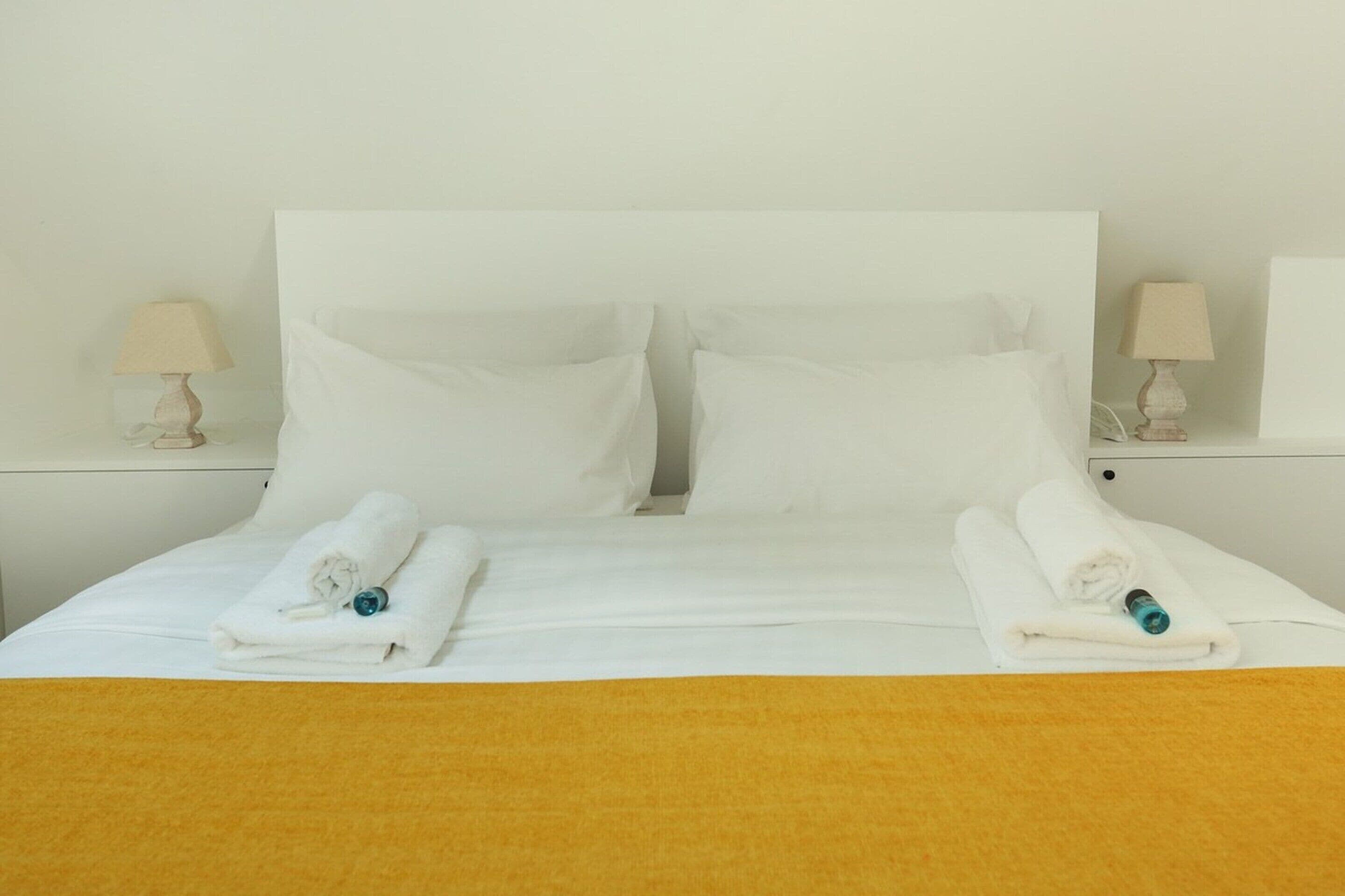 Camera d'albergo con un letto grande, due cuscini, due abat-jour e due asciugamani arrotolati con un oggetto blu sopra.
