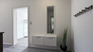 A hallway with a white wall, a mirror, a plant, and a wooden bench.