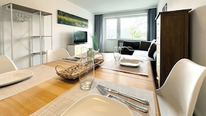 A modern dining area with a wooden table set for two, a television on a white cabinet, and a sofa in the background.