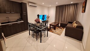 A modern living room with a sofa, a dining table with chairs, a TV, and a kitchen area with a sink and cabinets. - Loxwood House (Accra)