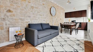 A modern living room with a stone wall, a grey sofa, a dining table with chairs, and a kitchen area in the background. - Welcoming Apartment - 2br/8p - Bonne-nouvelle (Paris)