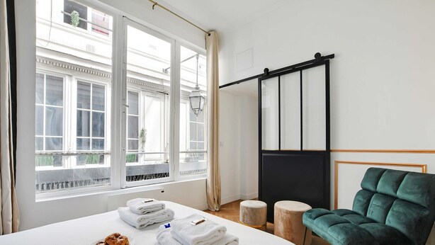 A hotel room with a bed, a green armchair, a small round table, and a view of the outside through large windows. - Welcoming Apartment - 2br/8p - Bonne-nouvelle (Paris)