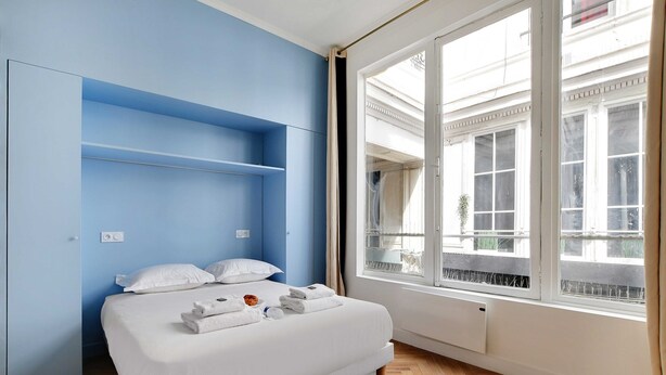 A bedroom with a bed, white bedding, a blue wall, a window with a view of buildings, and a radiator under the window. - Welcoming Apartment - 2br/8p - Bonne-nouvelle (Paris)
