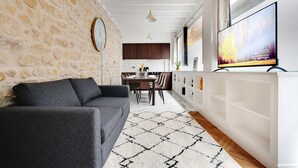 A modern living room with a stone wall, a grey sofa, a patterned rug, and a TV displaying a nature scene. - Welcoming Apartment - 2br/8p - Bonne-nouvelle (Paris)