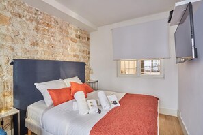 A hotel room with a stone wall, a bed with white bedding, orange pillows, a red blanket, a TV, and a window with blinds. - New Appartement Le Marais Center de Paris (Paris)