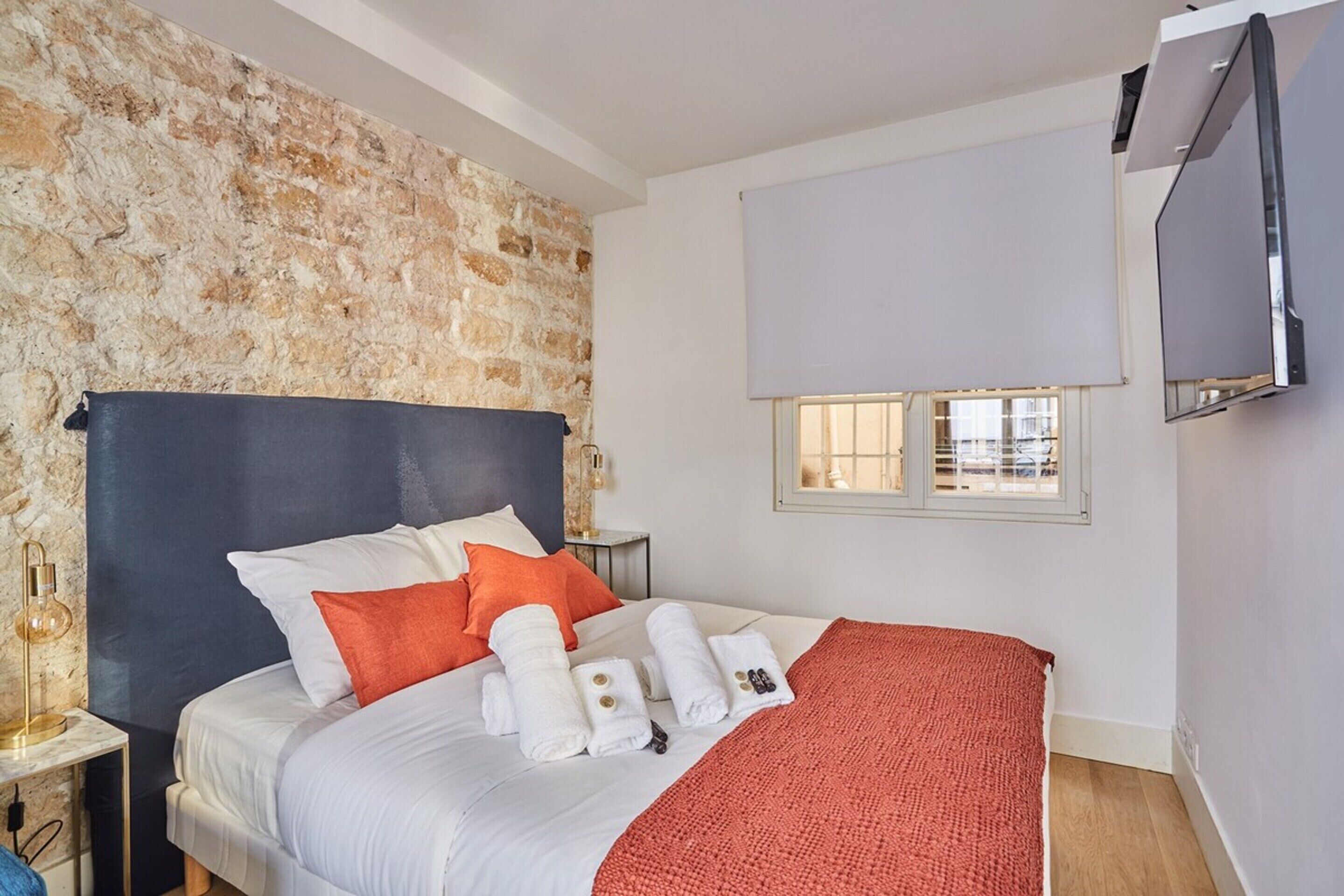 A hotel room with a stone wall, a bed with white bedding and orange pillows, a red blanket, and a TV mounted on the wall.