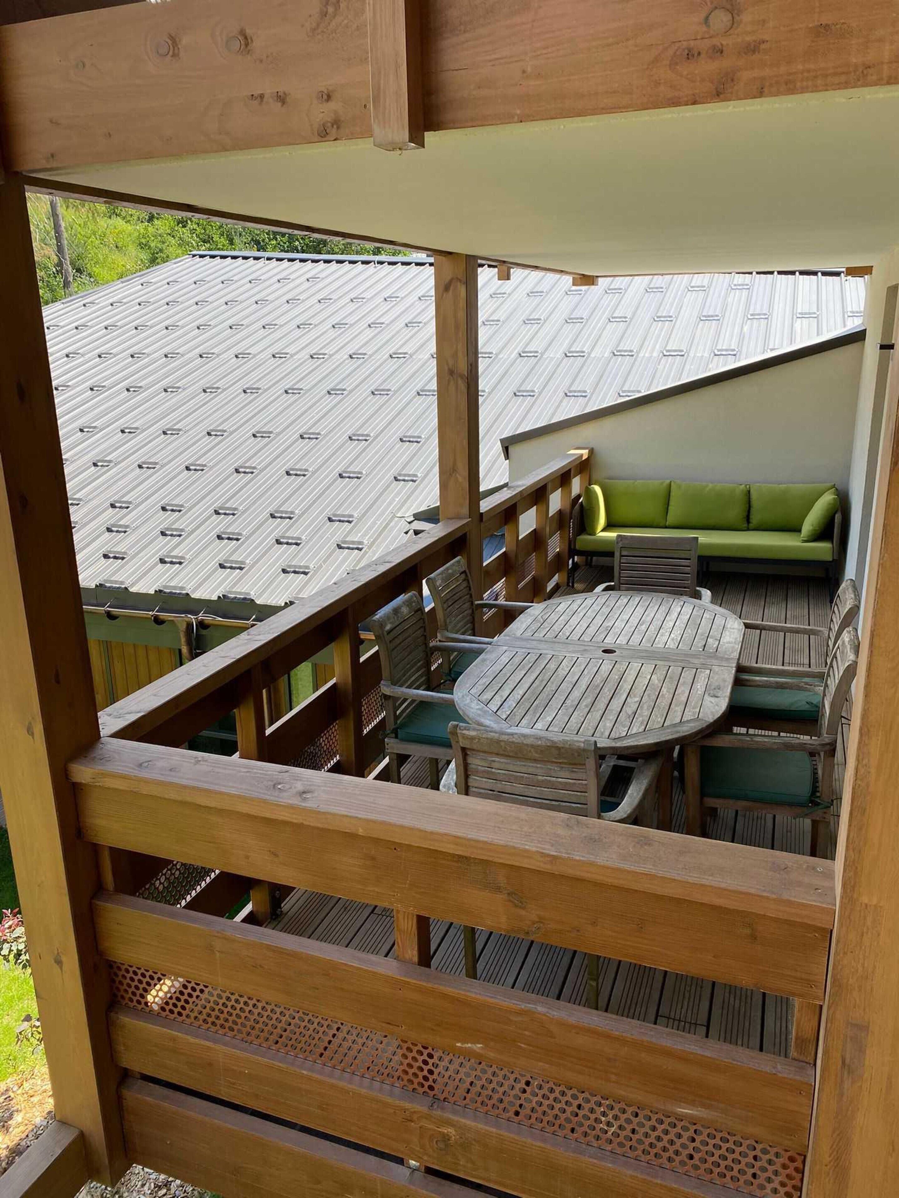 Eine Holzterrasse mit rundem Tisch und Stühlen, ein grünes Sofa und Blick auf ein Gebäude mit Metall-Dach.