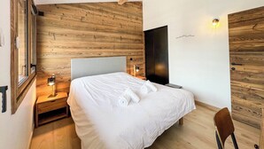 A modern bedroom with a wooden wall panel, a bed with white linens, a nightstand with a lamp, and a chair.