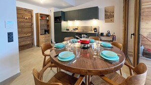 A dining area with a round wooden table set for six, surrounded by wooden chairs. A kitchen with dark cabinets and a wooden ceiling is visible in the background.