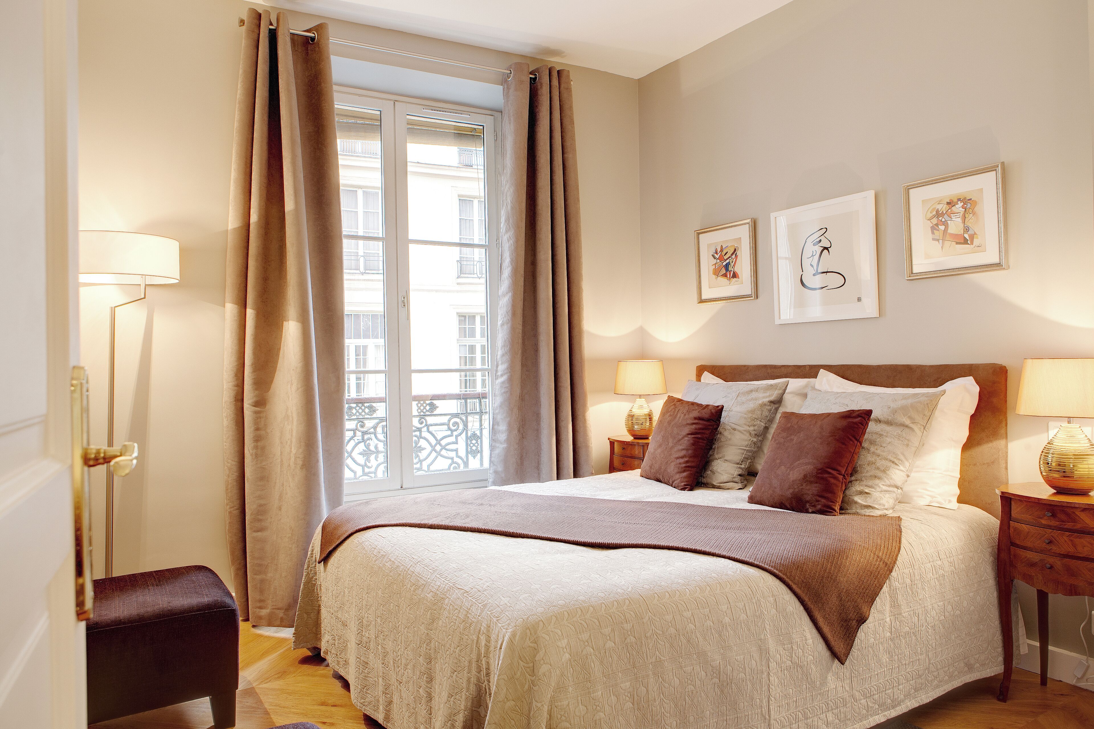 Une chambre à coucher avec un grand lit, deux tables de chevet avec des lampes, une fenêtre avec des rideaux et des œuvres d’art accrochées au mur.