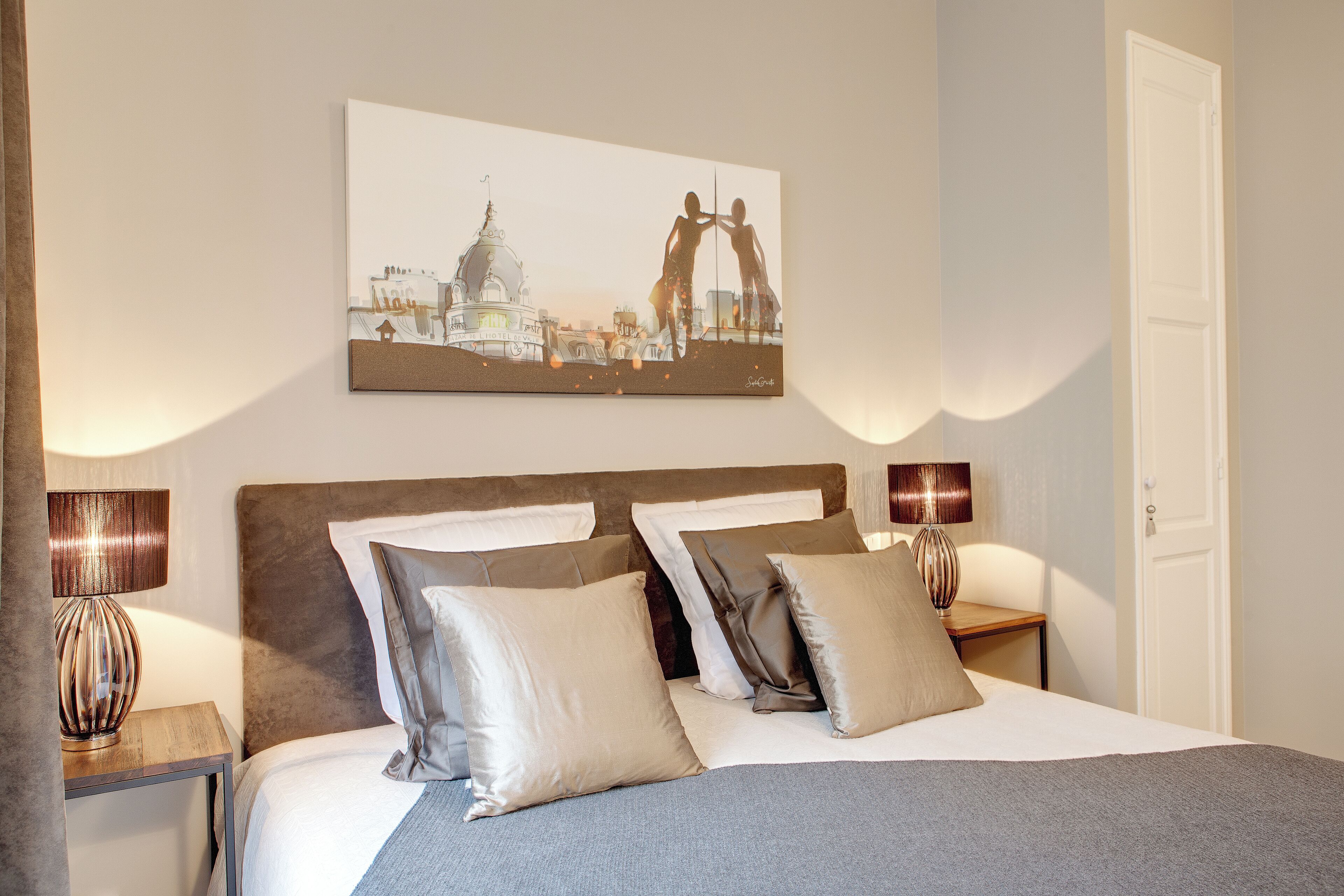 Un lit bien fait, avec une tête de lit, deux tables de chevet dotées de lampes et une œuvre d’art accrochée au mur représentant un paysage urbain.