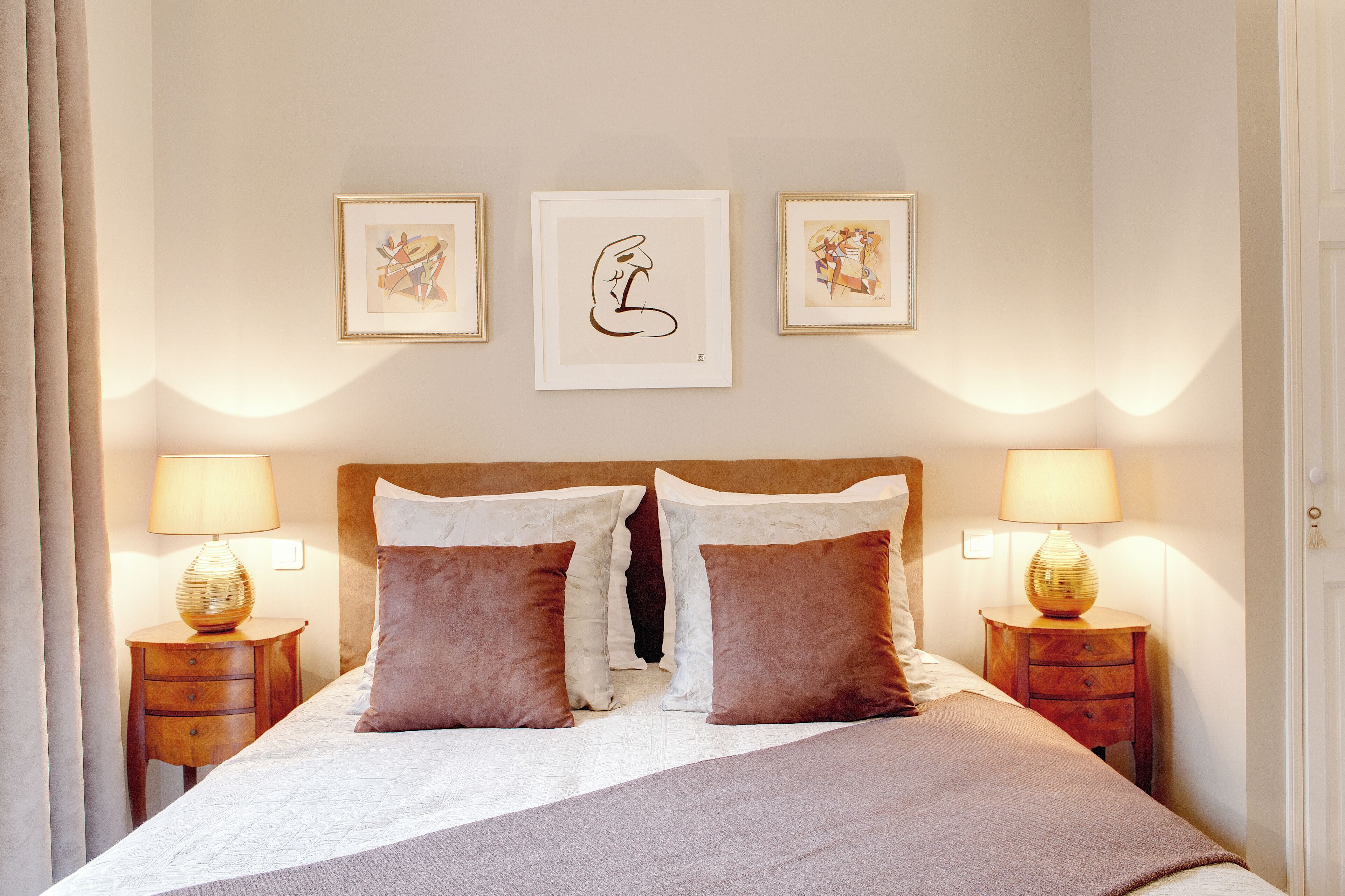 Een net opgemaakt bed met twee kussens, twee nachtkastjes met lampen, en drie ingelijste kunstwerken aan de muur.