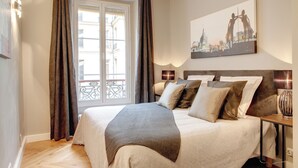 A neatly arranged bedroom with a large bed, a bedside table with a lamp, and a window with curtains.