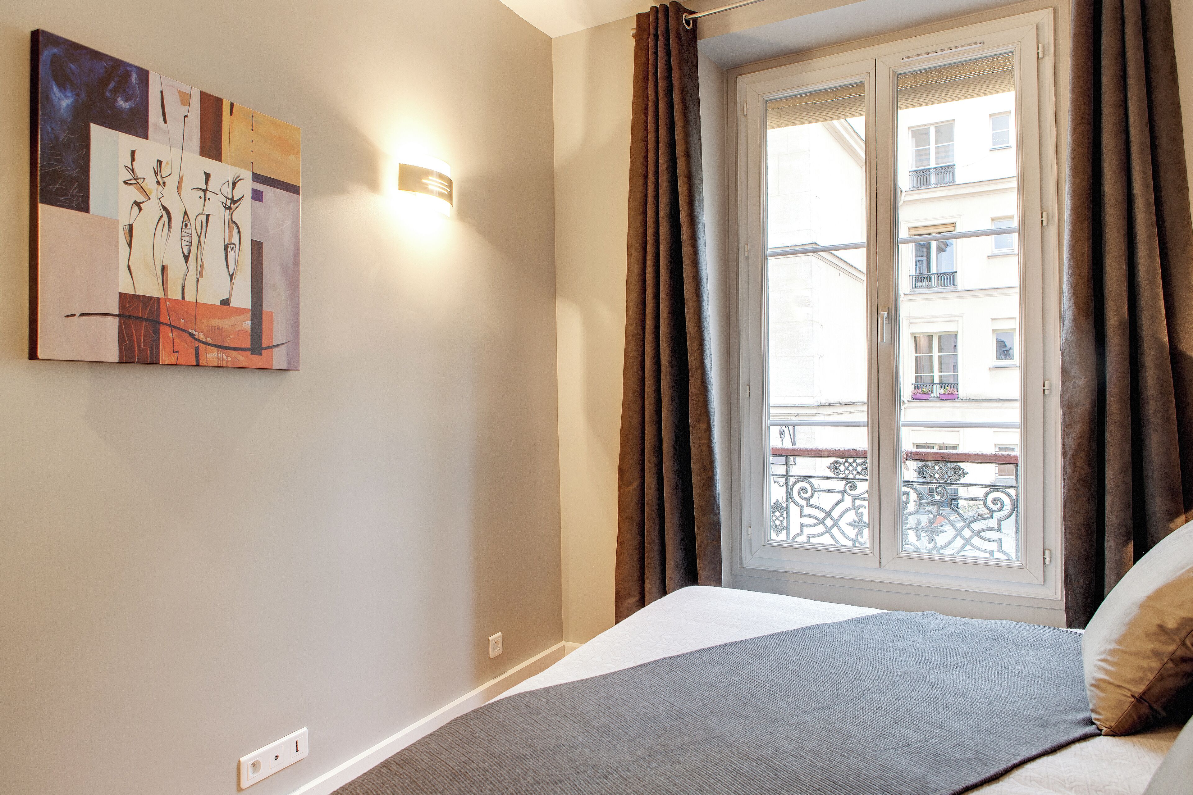 A hotel room with a bed, a window with curtains, a wall-mounted artwork, and a light fixture.
