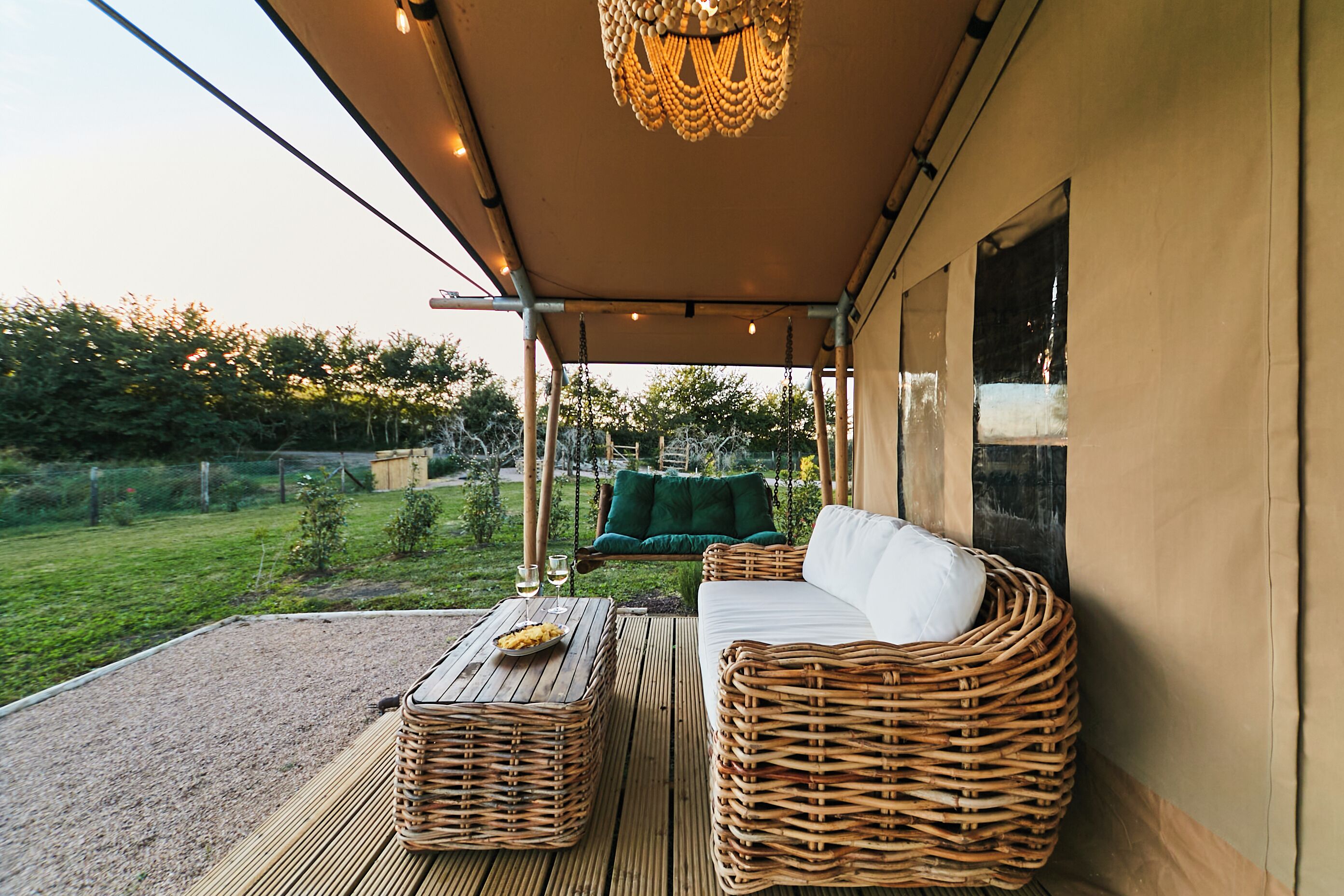 A covered outdoor seating area with wicker furniture, a hanging light fixture, and a view of a garden.