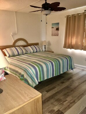 Ett sovrum med en säng i trä, randigt sänglinne, en takfläkt, ett nattduksbord, en lampa och ett fönster med gardiner.