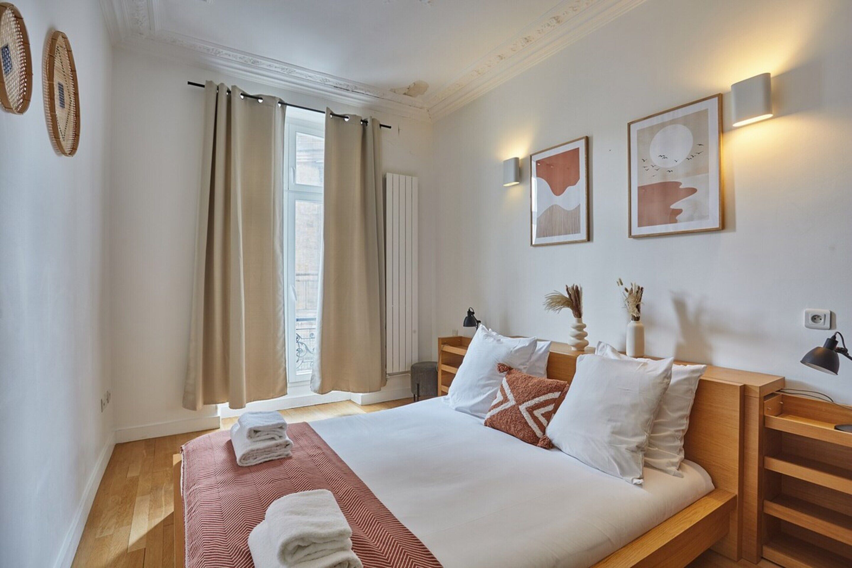 A neatly made bed with a striped bedspread, a wooden bedside table, and a nightstand with a lamp. There are two wall-mounted light fixtures and a framed picture on the wall.