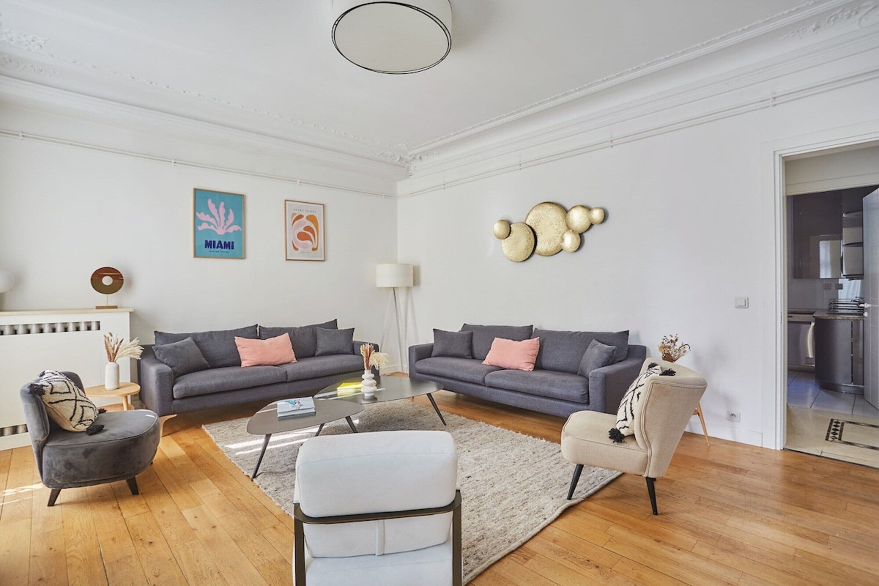 Ein geräumiges Wohnzimmer mit zwei grauen Sofas, einem weißen Sessel und einem Couchtisch. An den Wänden befinden sich gerahmte Bilder und ein dekoratives Wandelement.