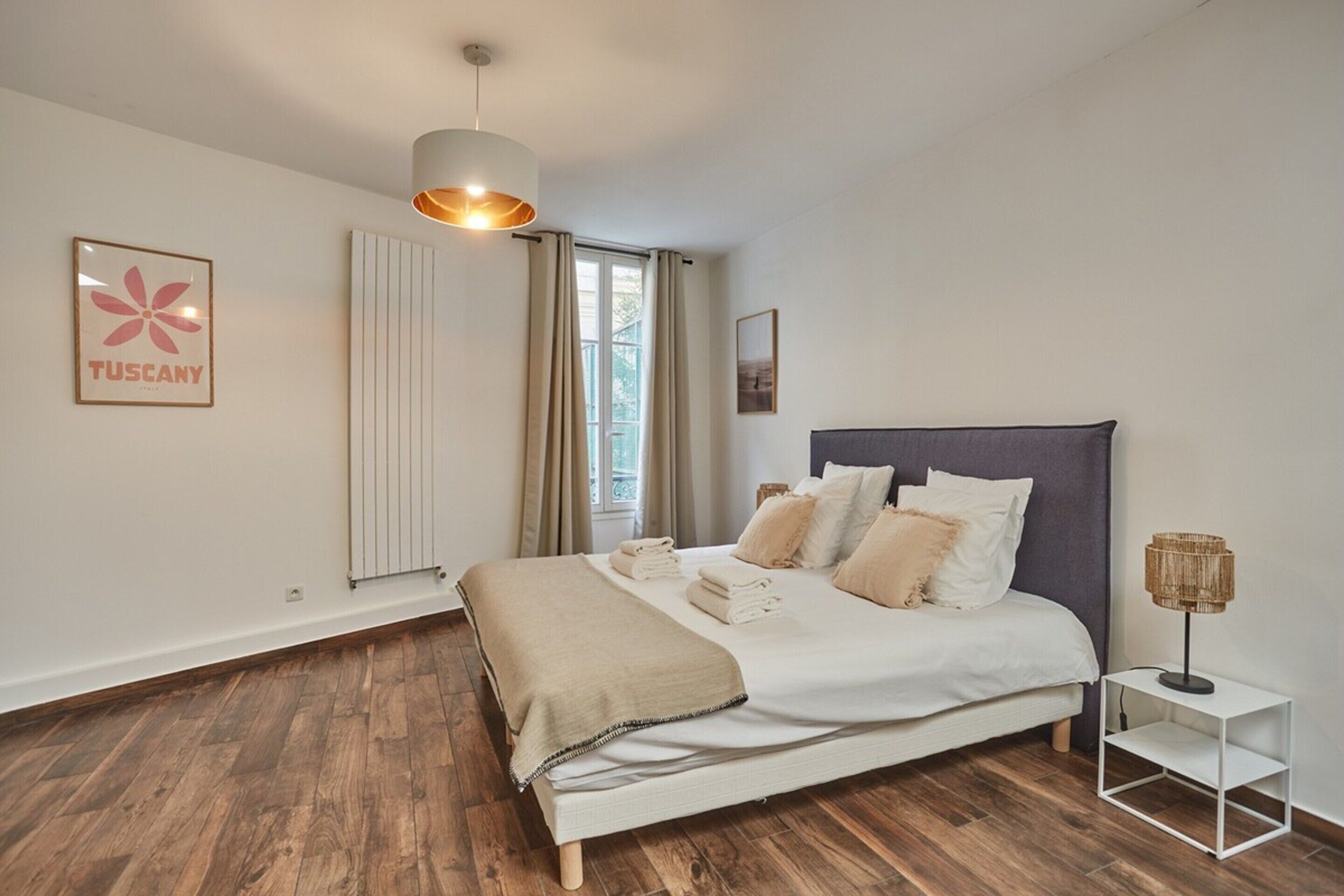 A bedroom with a bed, a bedside lamp, a radiator, a window with curtains, and a framed picture on the wall.
