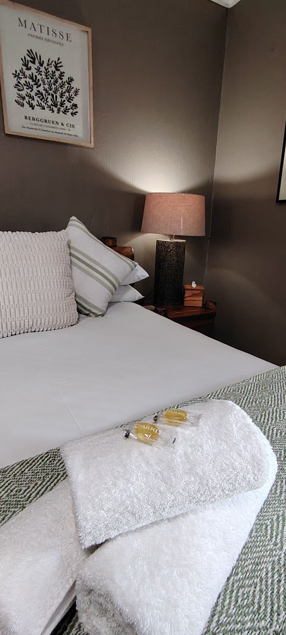 Een netjes opgemaakt bed met witte lakens, een kussen met groene en witte strepen, en een witte handdoek met gele bloemen. Daarboven hangt aan de muur een lijst met een plant.