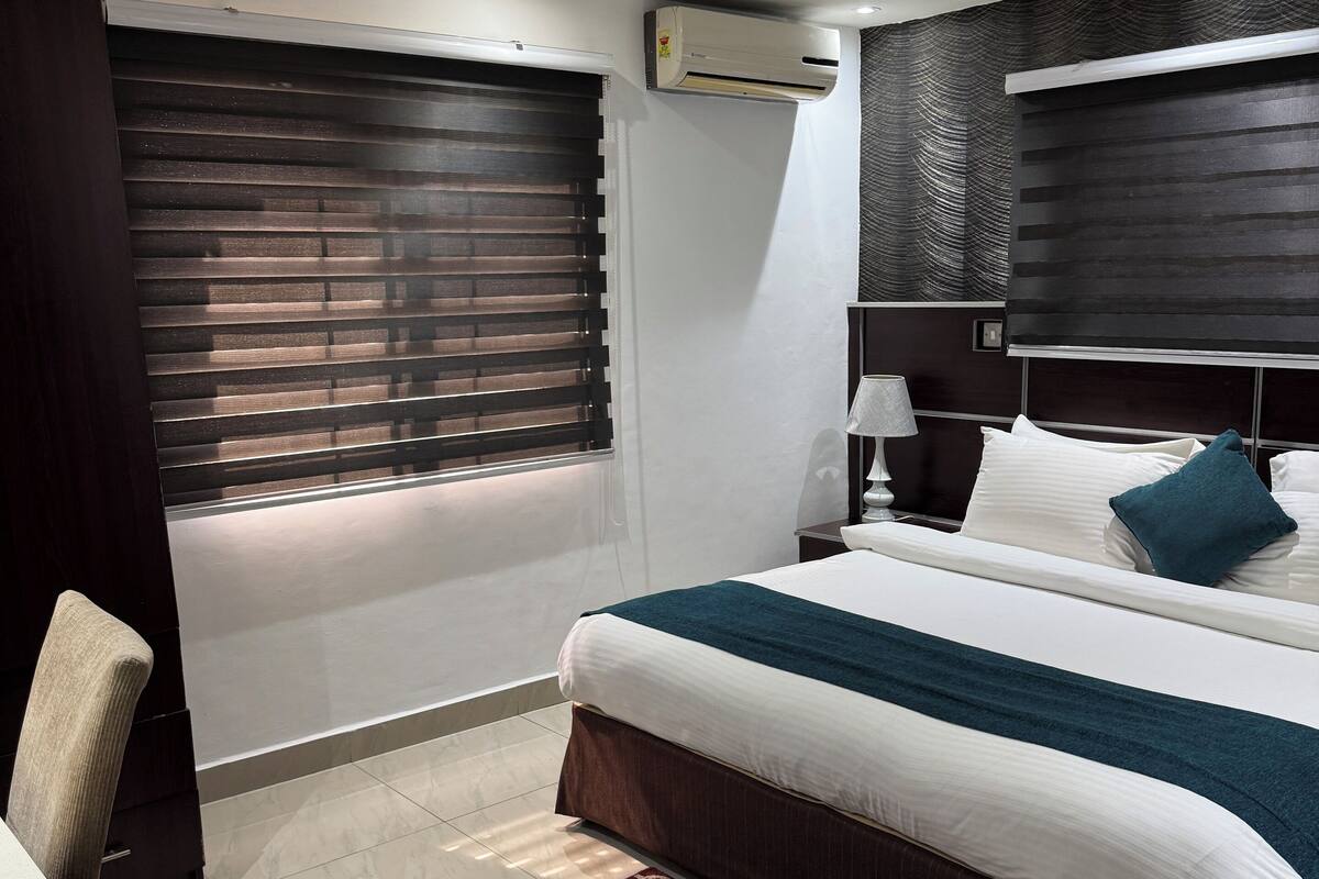 A hotel room with a bed, bedside table, lamp, air conditioning unit, window with blinds, and a patterned rug.