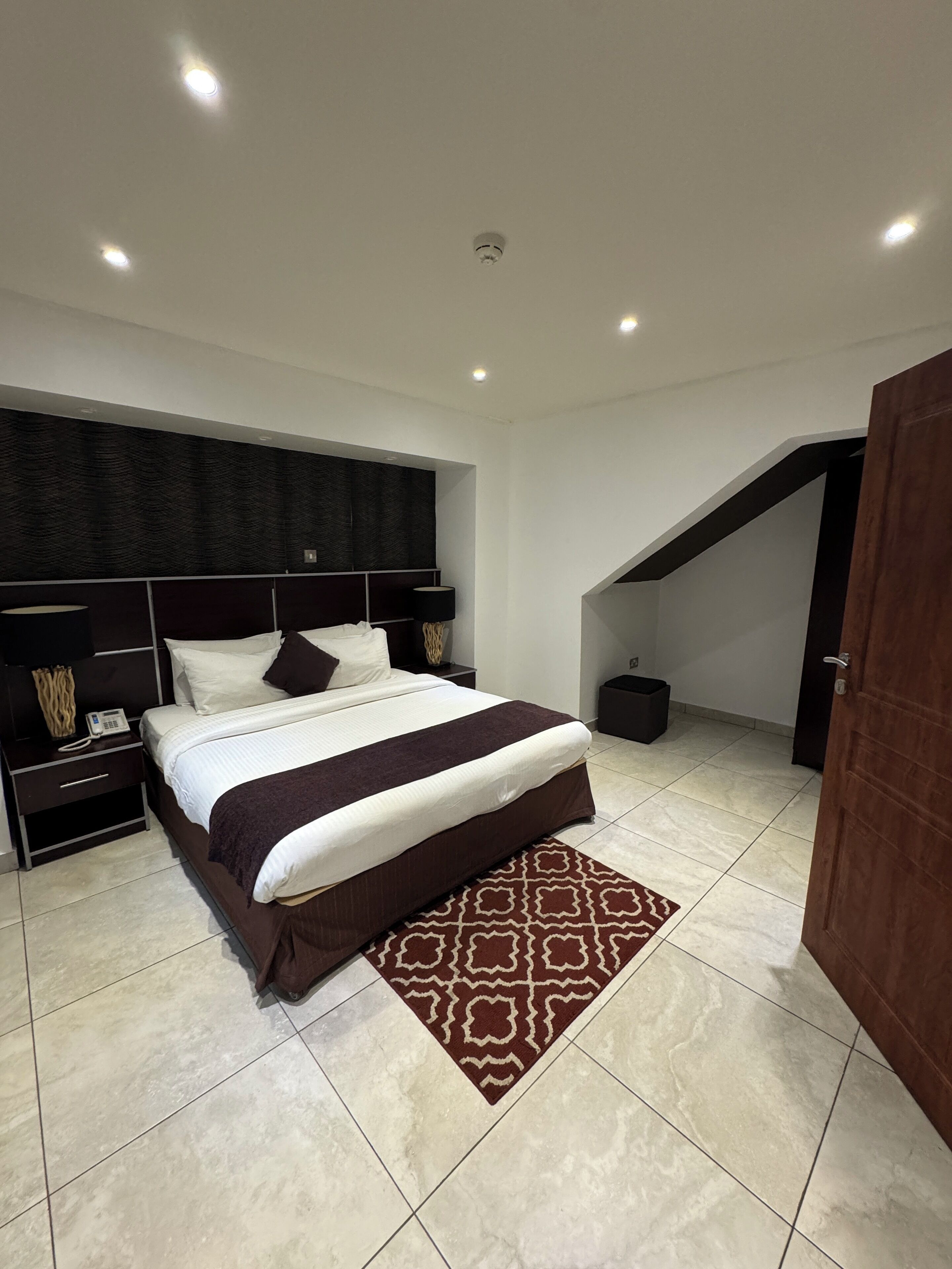 Quarto moderno com cama grande, mesas de cabeceira, tapete estampado e escada para um andar superior.