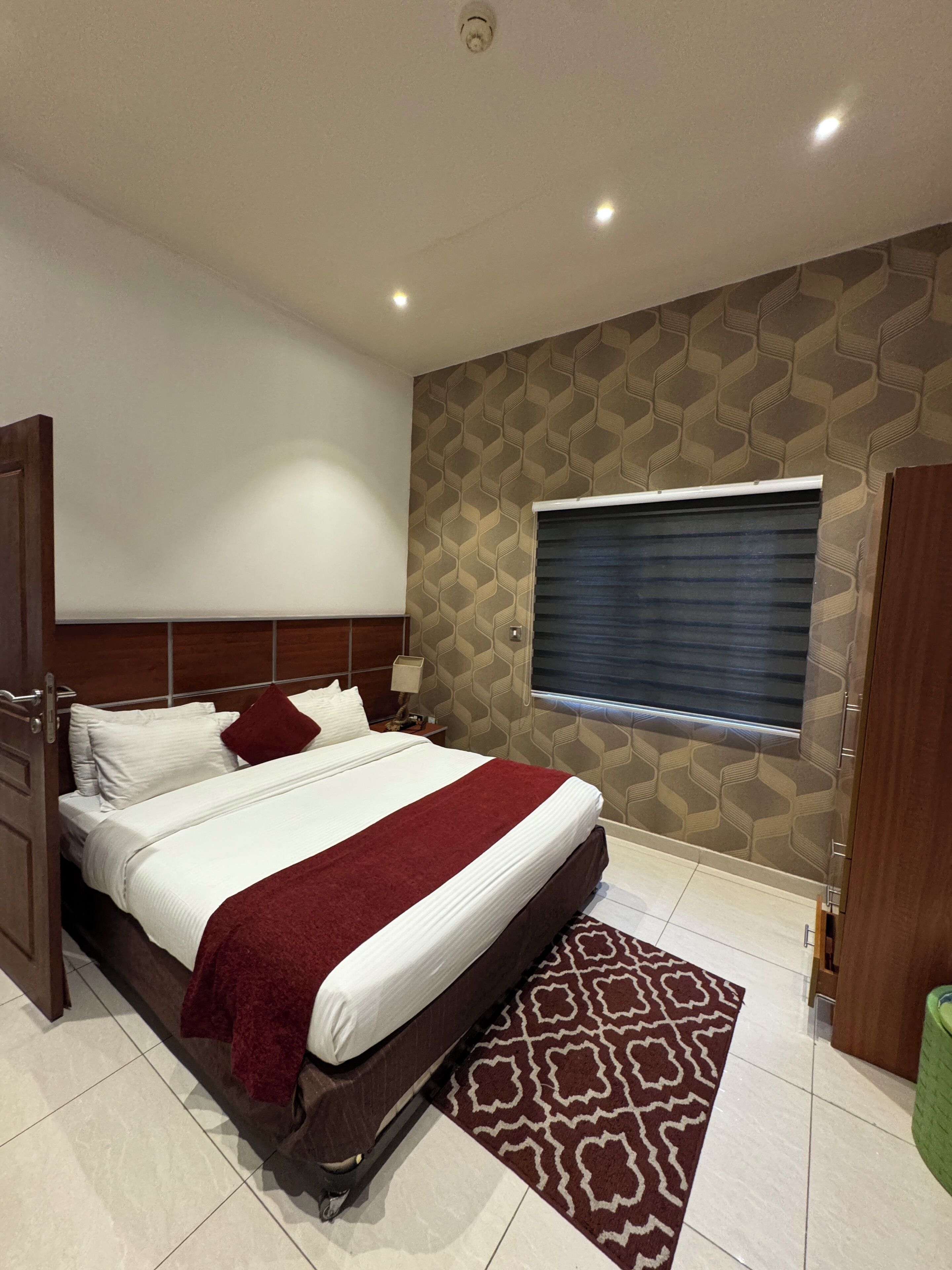 Quarto de hotel moderno com uma cama grande, parede estampada, janela com persianas e uma lixeira verde.