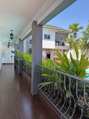 A covered balcony with a metal railing, potted plants, and a view of a residential building with a pool. - Eagles Accra (Accra)