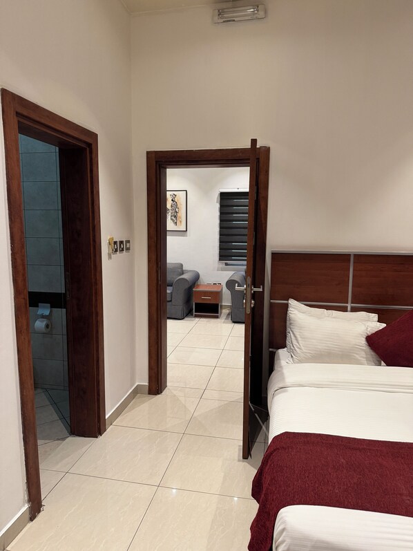 A hotel room with a bed, a small bedside table, a chair, and a view of the hallway. - Eagles Accra (Accra)