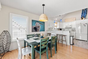 A modern kitchen with white cabinets, a dining table with chairs, and a large painting of a bird. - Coral Sunset: Private Pool | Gated Golf Community (Corolla)