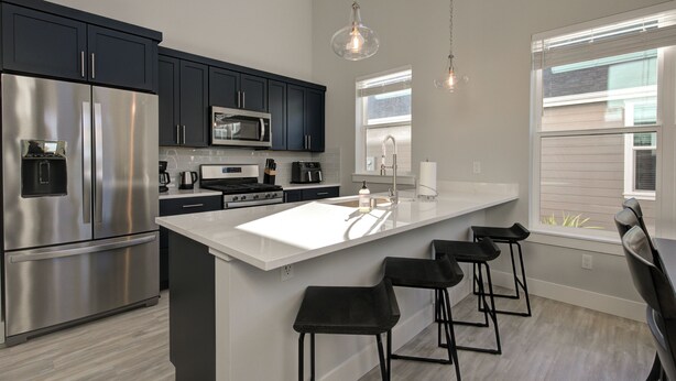 A modern kitchen with stainless steel appliances, dark cabinets, a white island with bar stools, and pendant lighting. - Welcome to your ultimate getaway, Burl by the Bay! (Ocean Shores)