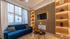 A modern living room with a blue sofa, a flat-screen TV mounted on a wooden panel, and a small table with a lamp and decorative items. - Apartment Centre de Paris Sacre Coeur (Paris)