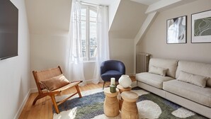 A modern living room with a wooden chair, a blue armchair, a beige sofa, and a large window with sheer curtains. - Superb Apartment Arc de Triomphe/ Trocadero (Paris)