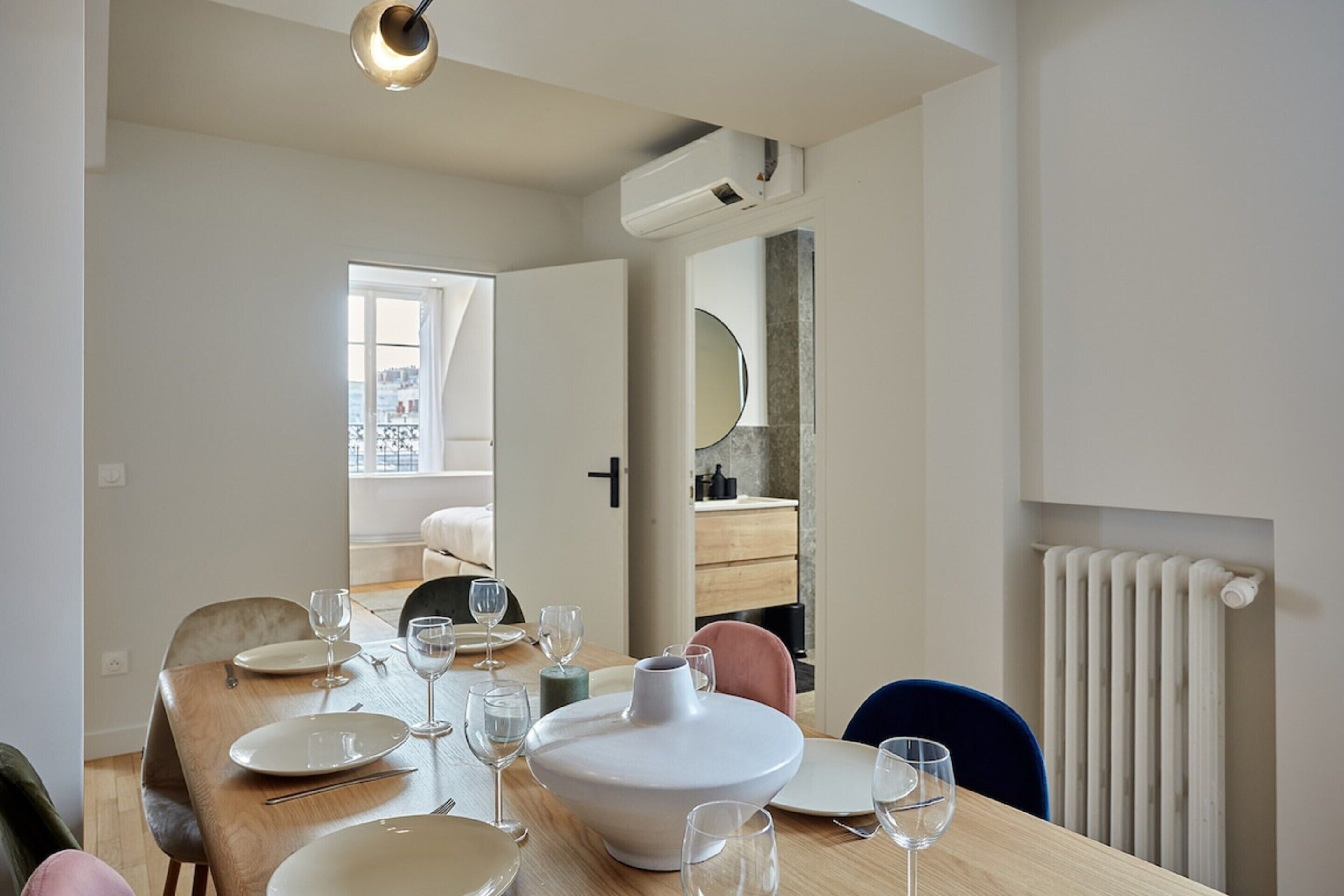 A dining area with a wooden table set for four, a large white vase, and a view of a cityscape through an open door.