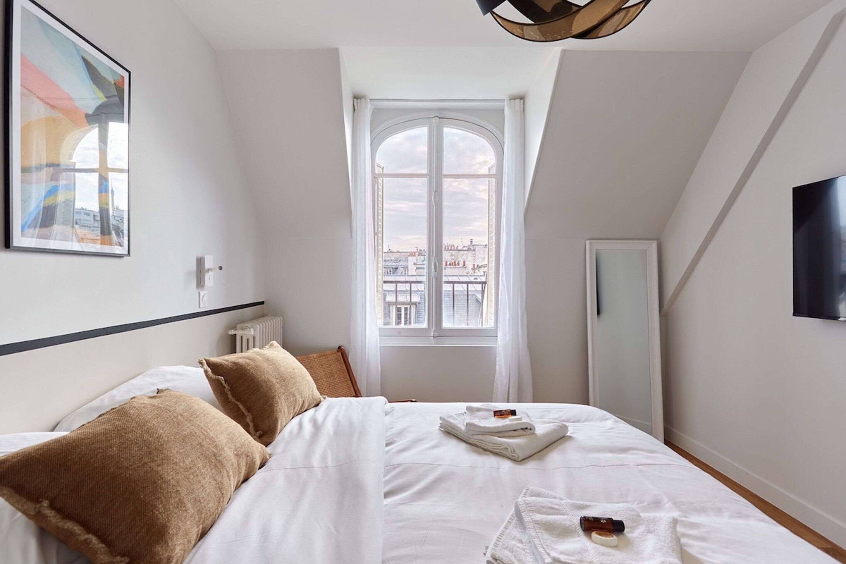 A bedroom with a large bed, a window with a view of buildings, a radiator, and a flat-screen TV on the wall.
