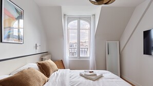 A bedroom with a large bed, a window with a view of buildings, a radiator, and a flat-screen TV on the wall. - Superb Apartment Arc de Triomphe/ Trocadero (Paris)