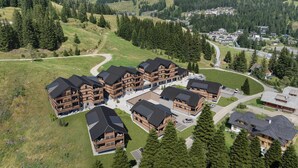 Eine Ansammlung von blockhausartigen Gebäuden mit mehreren Stockwerken, gruppiert um einen Innenhof, gelegen in einer gebirgigen Gegend mit dichten Wäldern und einer kurvenreichen Straße.
