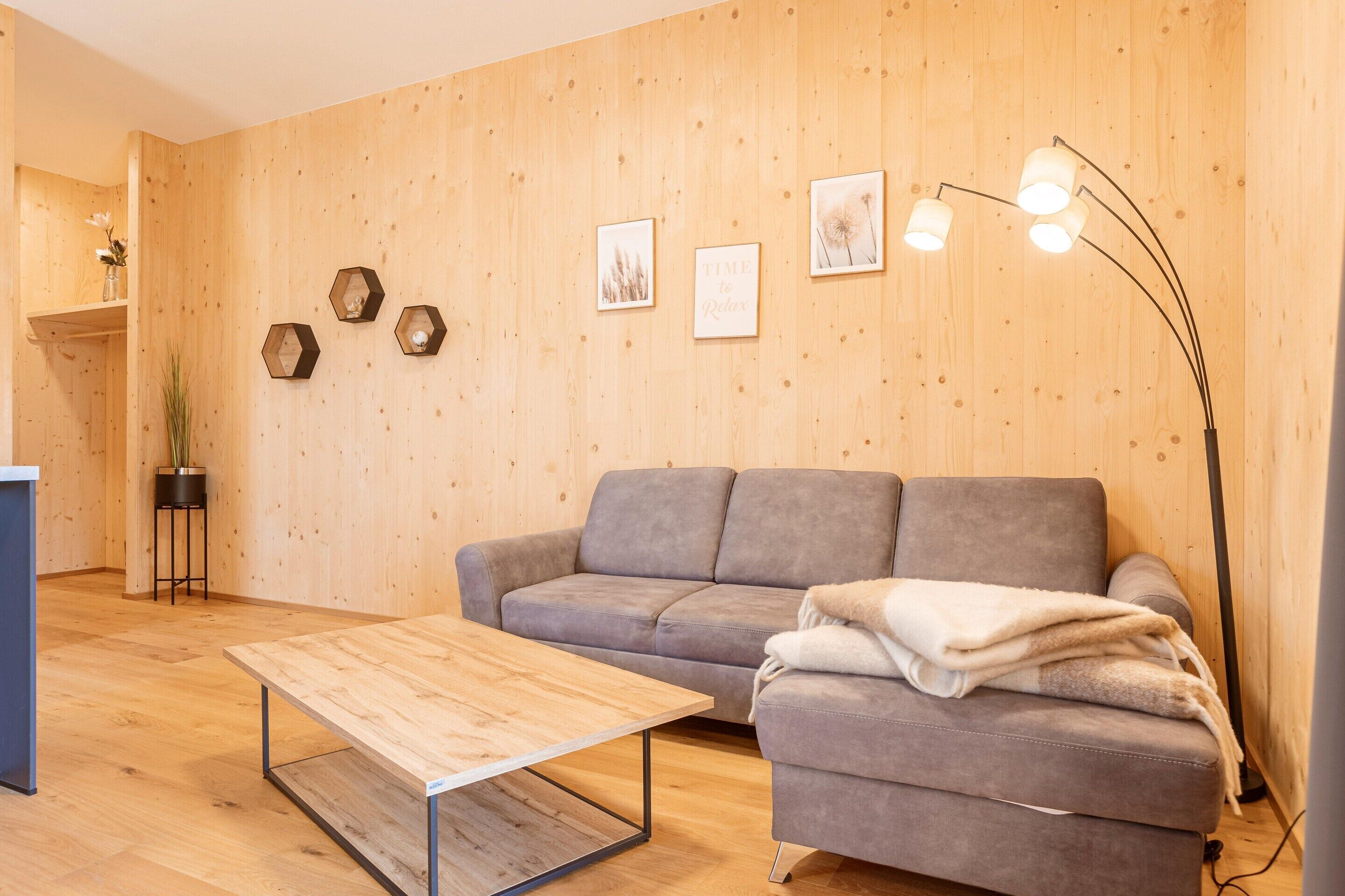 Ein modernes Wohnzimmer mit einem grauen Sofa, einem Holztisch und einer Stehlampe. Es gibt sechseckige Wanddekorationen und eine kleine Pflanze auf einem Sockel.