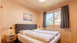A bedroom with wooden walls, a bed with white linens, a nightstand with drawers, a lamp, and a window with curtains. - Ferienwohnung mit 2 Schlafzimmern & Sauna (Oberwölz)