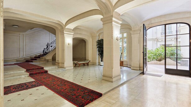 Un hall d’entrée spacieux avec de hauts plafonds, un grand escalier et un tapis rouge menant à une porte.