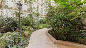 Un sentier étroit et sinueux, bordé d’une végétation luxuriante, avec un bâtiment blanc doté de balcons.
