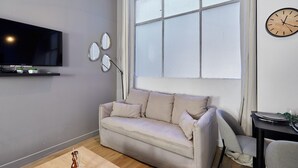 A living room with a sofa, a flat-screen TV, a window with blinds, a clock, and a small table with a chair. - Suite Saint Germain des Pres Rue de Rennes (Paris)