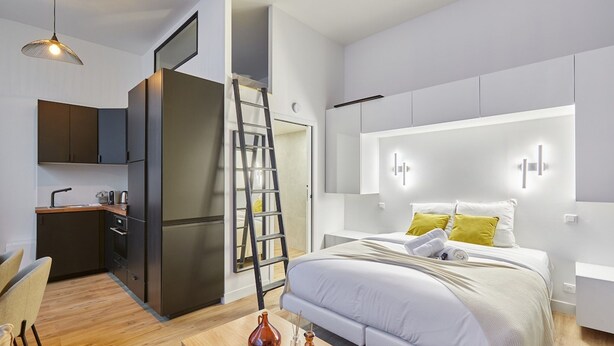 A modern bedroom with a large bed, bedside tables, a desk, and a kitchen area with cabinets and a refrigerator. - Suite Saint Germain des Pres Rue de Rennes (Paris)