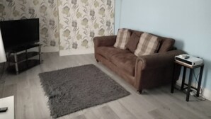 A living room with a brown sofa, a grey rug, a television on a stand, and a small table with a projector. - Sunny Seaside Apartment in Blackpool (Blackpool)