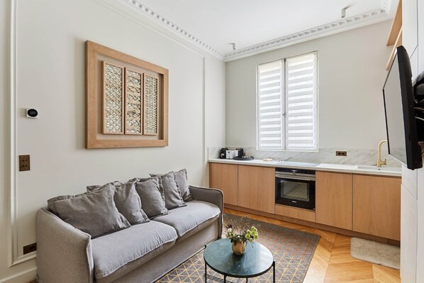 A modern apartment with a grey sofa, a small round table with a vase, a kitchen with wooden cabinets, and a large window with white shutters. - Magnificent Apartment Etoile (Paris)