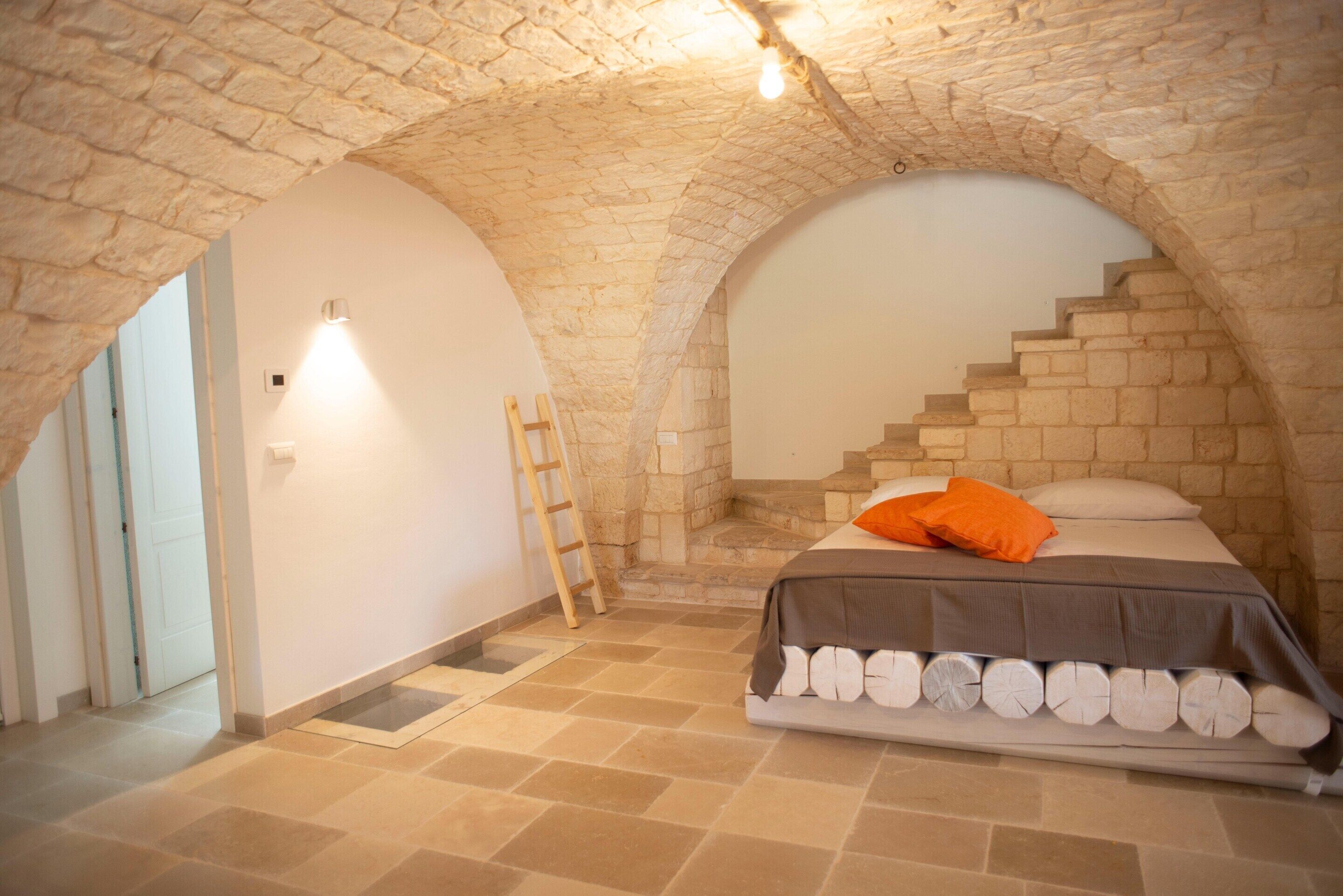 A bedroom with a stone vaulted ceiling, a bed with a white mattress and orange pillow, and a wooden ladder against the wall.