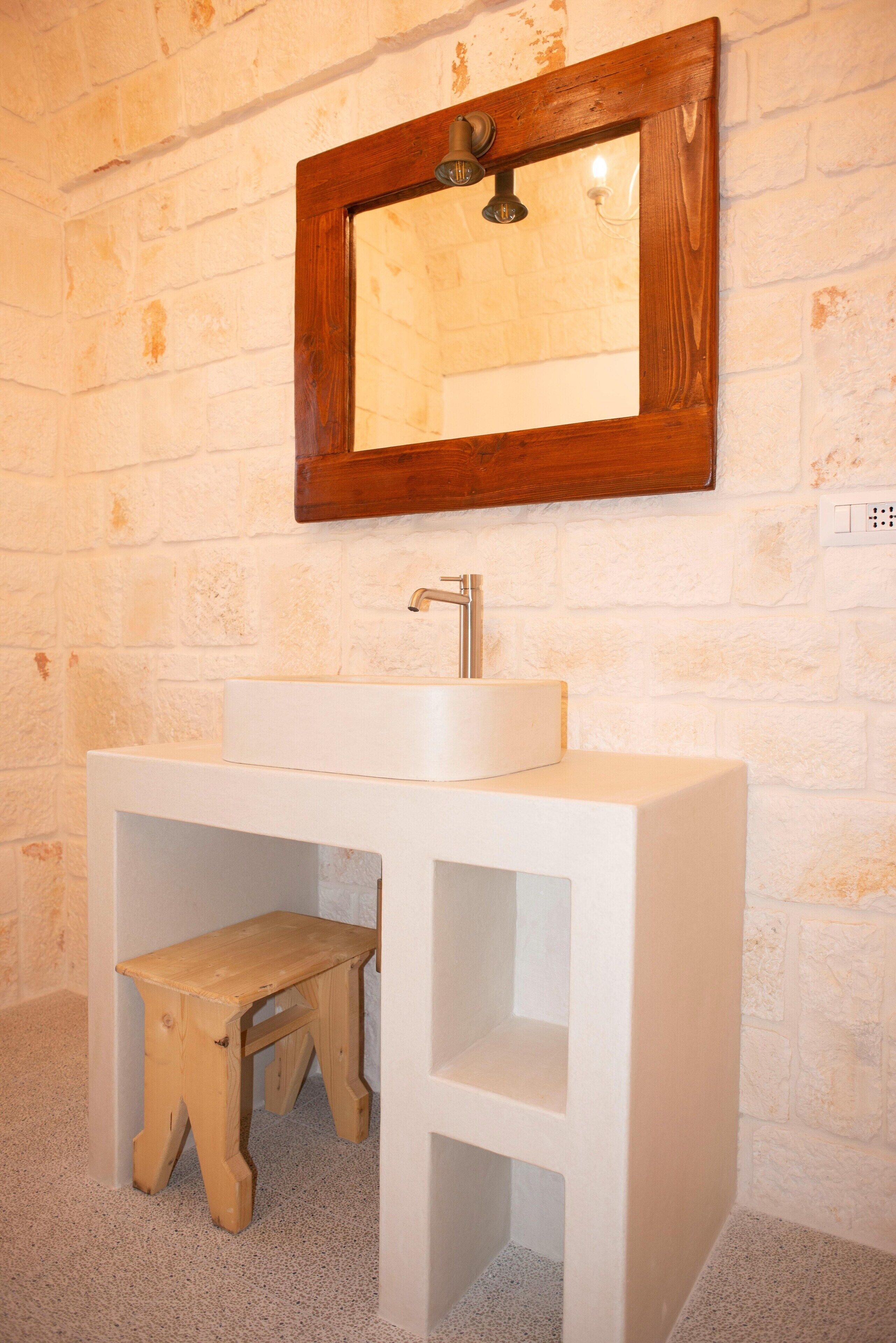 Un baño con lavamanos blanco, un taburete de madera y un espejo con marco de madera.